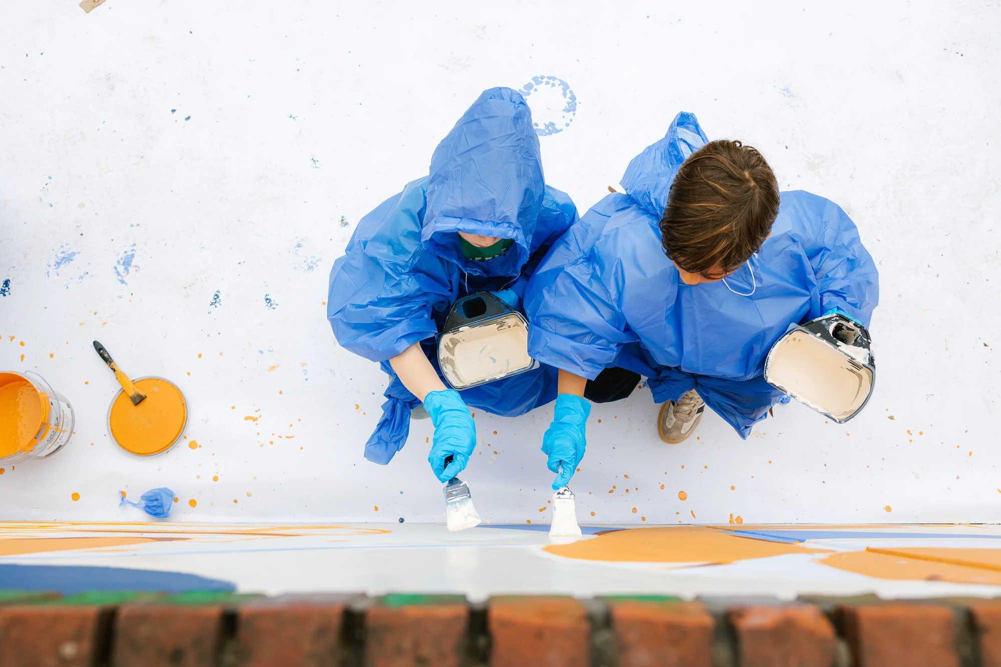 Twee kinderen in blauwe regenjassen schilderen van bovenaf gezien met kwasten op een muurdeel in blauw en oranje