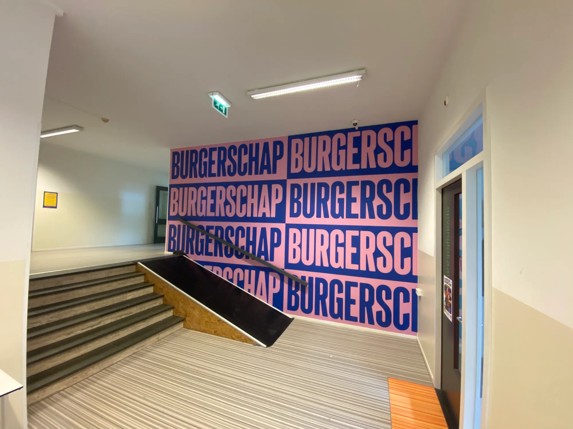 Wand in een schoolgebouw van Curio met het woord BURGERSCHAP herhaald in roze letters op donkerblauw
