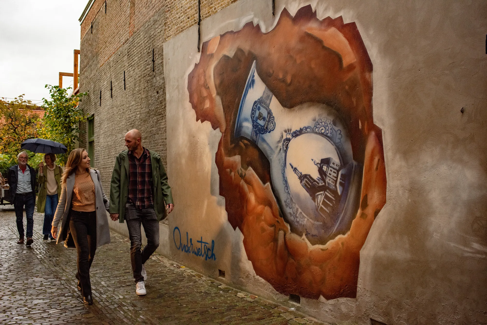 Muurschildering voor restaurant Ouderwetsch: een Delfts blauw bord dat door een roestbruine muur heen breekt, voorbijgangers met paraplu